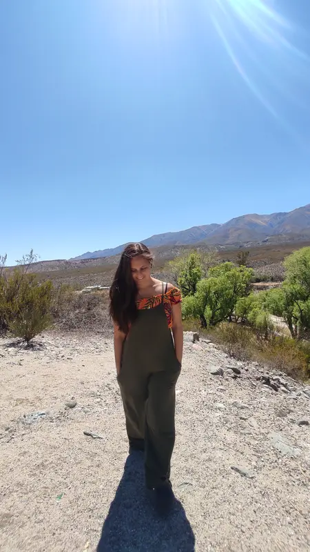 Andrea con un enterito verde, sonriendo con la mirada baja en un camino de tierra entre montañas, bajo un cielo despejado