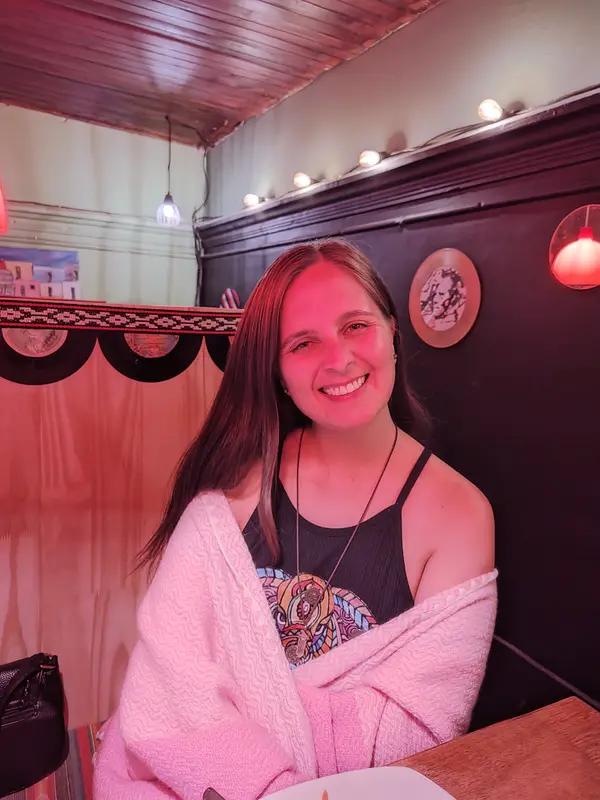Andrea sonriendo sentada a una mesa de un bar cálido, con un abrigo rosa sobre los hombros