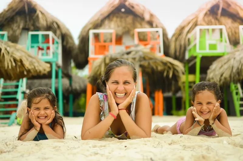 Andrea Cazaux y sus mellizas sonriendo recostadas en la arena, frente a cabañas de techo de paja en la playa
