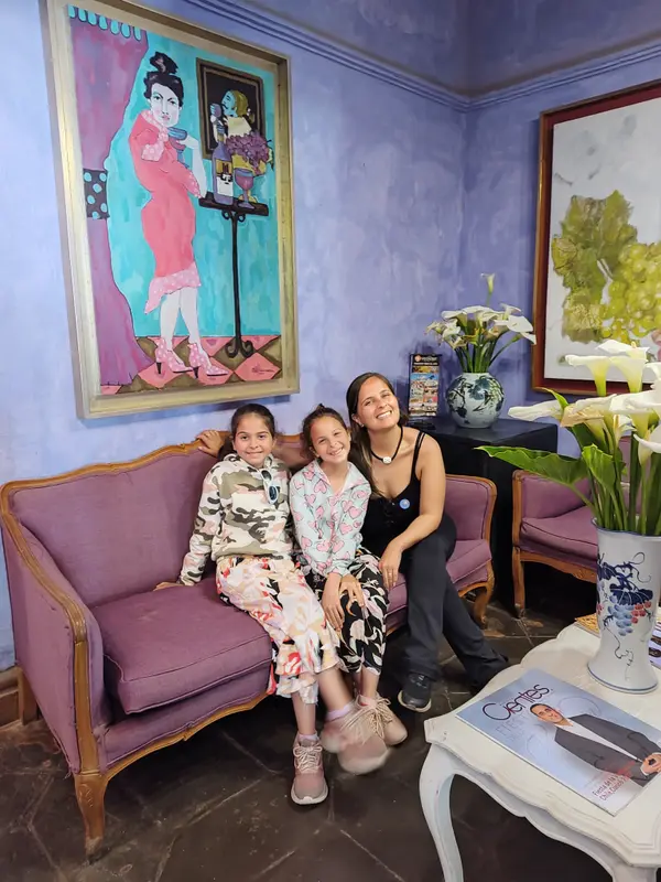 Andrea y sus mellizas sonriendo juntas en un sillón violeta, bajo un cuadro colorido en una sala de paredes lilas