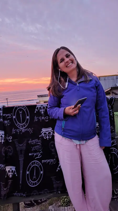 Andrea con buzo azul y celular en la mano, frente al mar durante un atardecer rosado