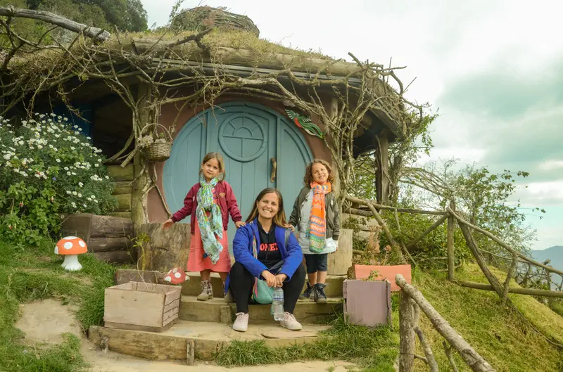 Andrea y sus mellizas sentadas en la puerta redonda de una casa estilo hobbit con techo de pasto