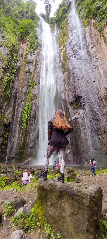 Andrea de espaldas frente a una cascada alta que cae entre paredes de roca cubiertas de vegetación