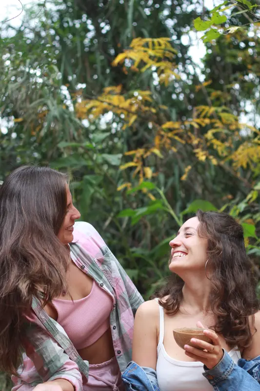 Andrea conversando y riendo con otra mujer al aire libre, entre árboles