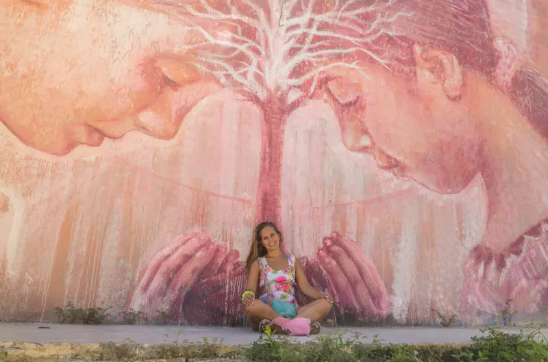 Andrea sentada al pie de un gran mural rosado con dos rostros y un árbol entre las manos