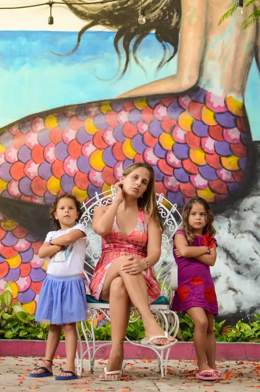 Andrea sentada en una silla blanca junto a sus dos hijas, delante de un mural colorido de una sirena