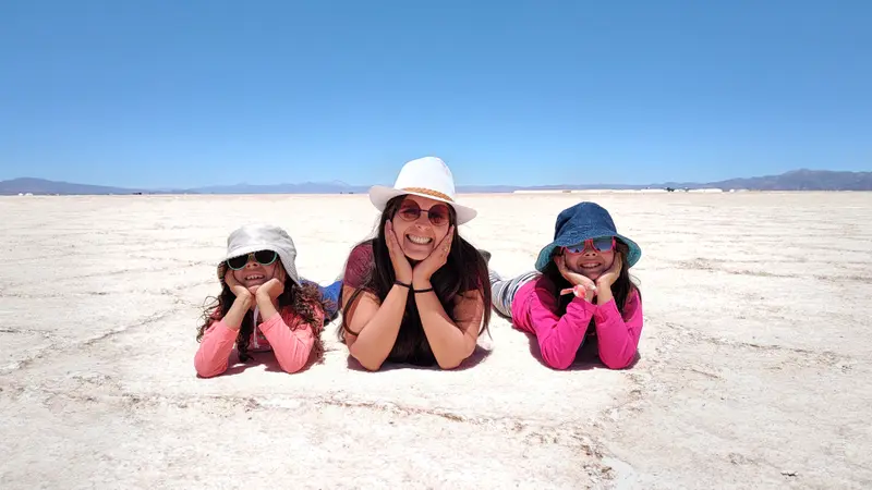 Andrea y sus dos hijas recostadas sobre la sal blanca de un salar, con sombreros y bajo un cielo azul