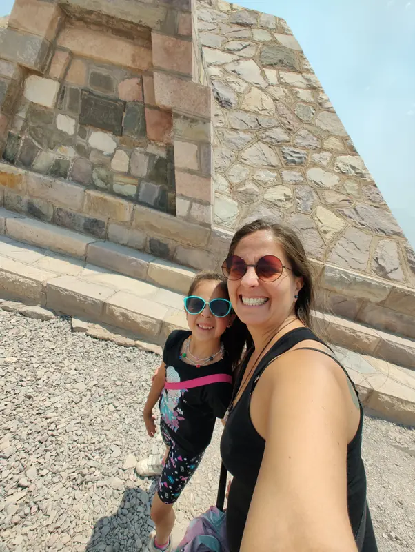 Andrea y una de sus mellizas, ambas con anteojos de sol, sonriendo en una selfie junto a un monumento de piedra