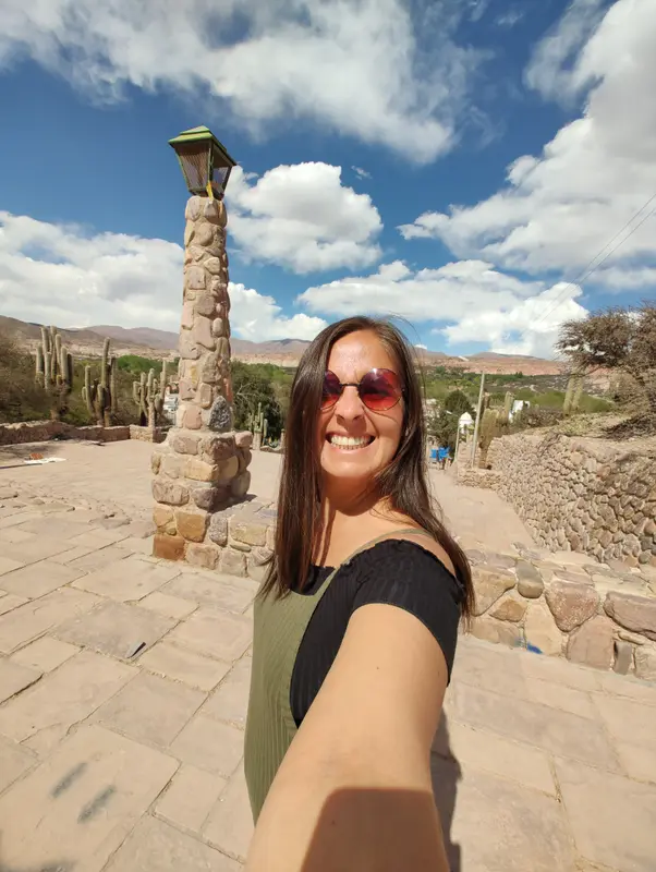 Andrea con anteojos de sol en una selfie en la plaza de piedra de un pueblo de montaña rodeado de cardones