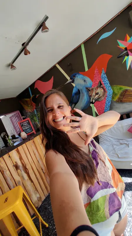 Andrea riendo en una selfie, saludando con la mano frente a un mural colorido de aves y un zorro